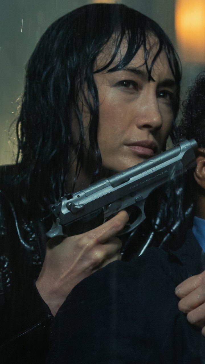 Phone wallpaper featuring Maggie Q in a tense scene from the movie The Protégé, holding a gun with Ray Fearon partially visible. Compatible with iPhones and Android devices.