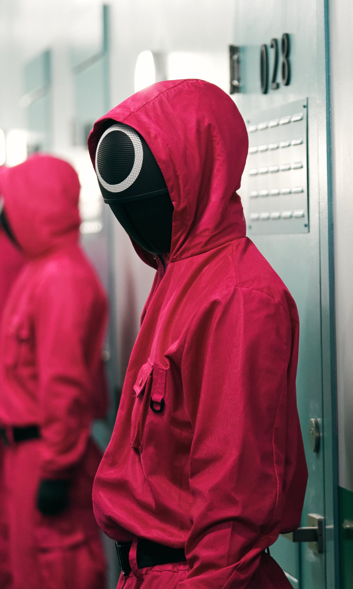 Phone wallpaper featuring a Squid Game TV show guard: masked figure in red hooded jumpsuit with circular black face mask standing by hallway lockers.