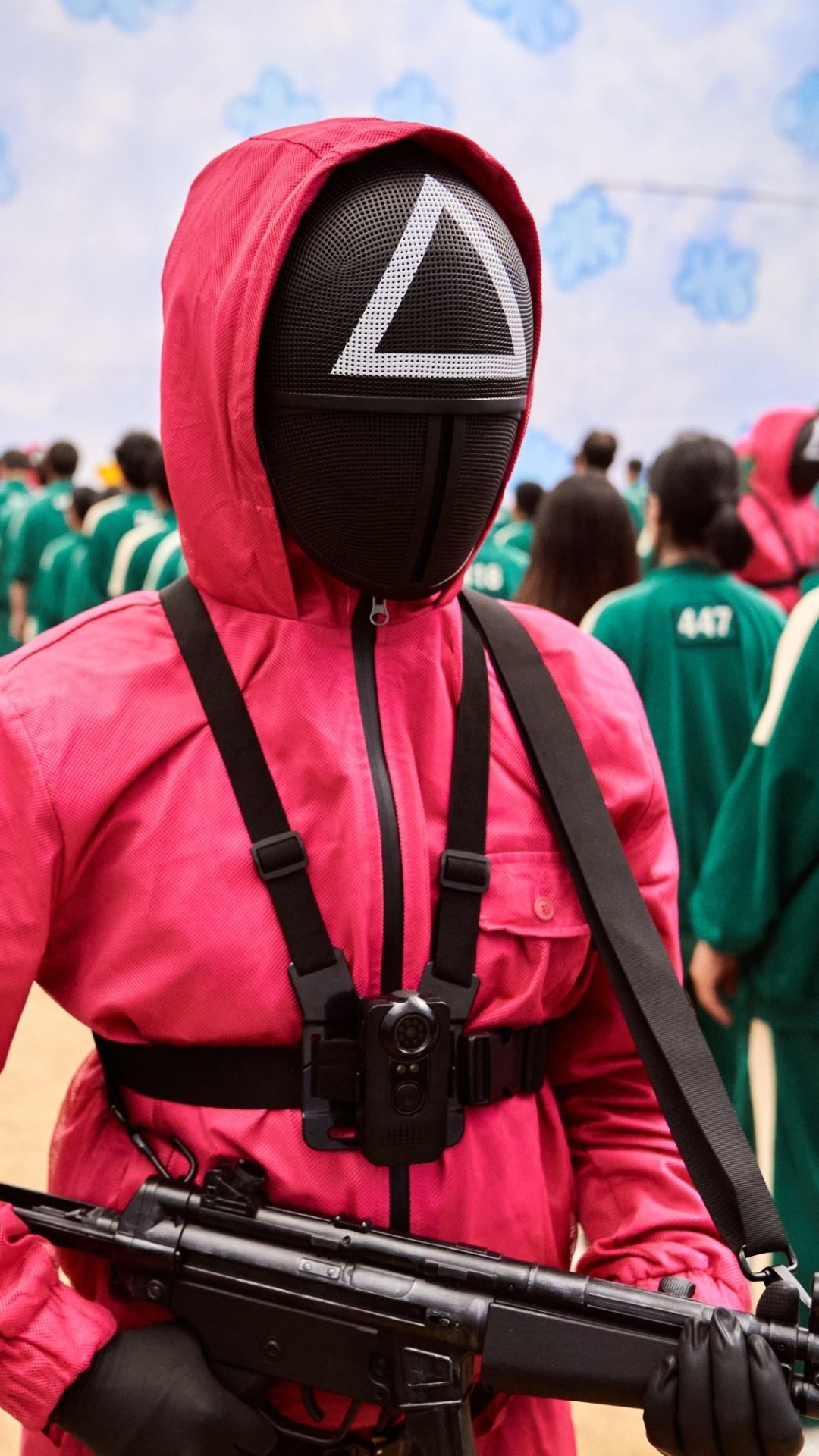 Phone wallpaper of TV show Squid Game: a masked guard in a pink suit with a triangle symbol holding a rifle, blurred players in green tracksuits in the background.