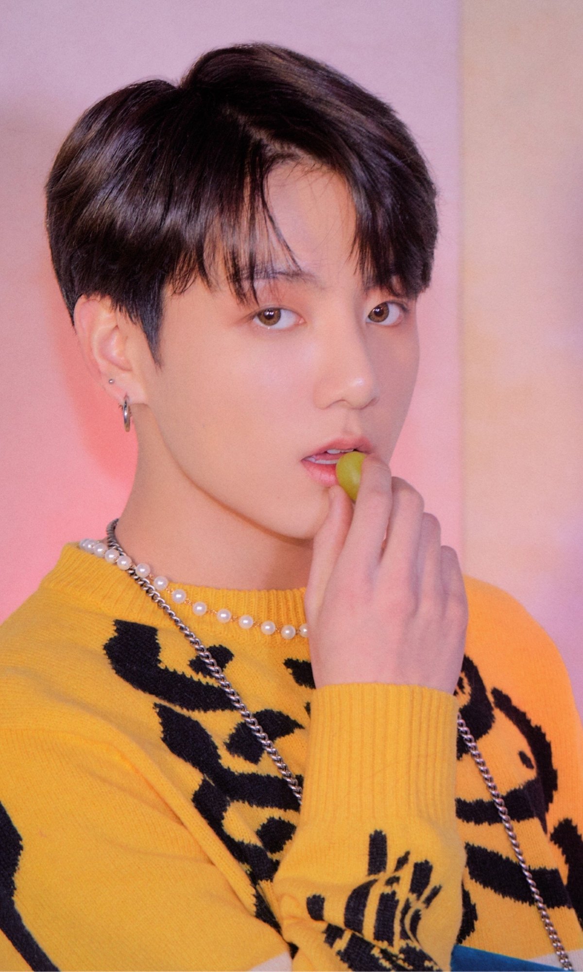 Phone wallpaper portrait of a K-pop singer: young man in a yellow sweater with chains, holding a green grape against a soft pink, BTS-inspired music aesthetic.