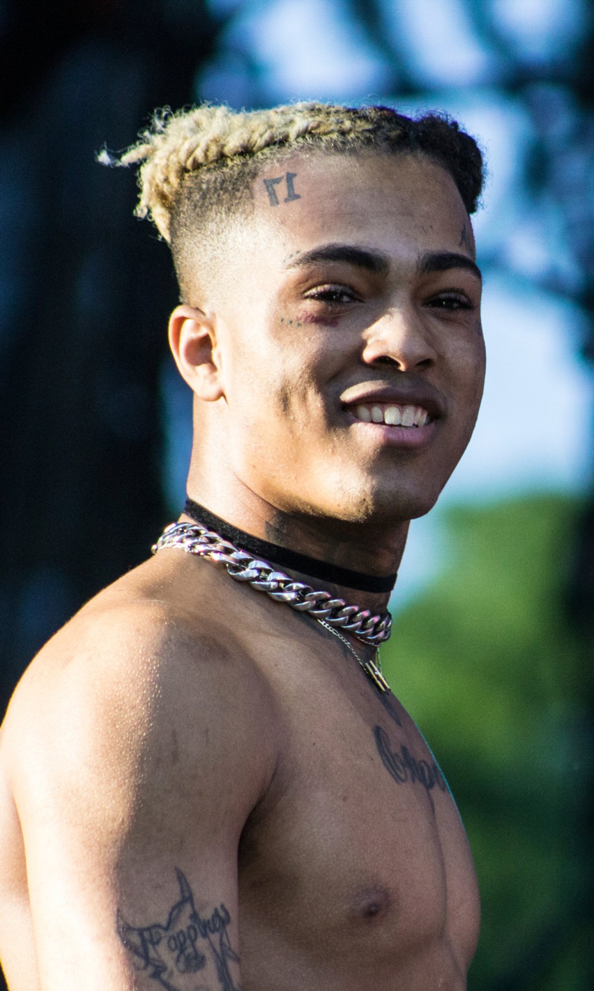 Phone wallpaper featuring a smiling, shirtless XXXTentacion with blonde hair, tattoos, and a chain necklace, set against a blurred outdoor background.