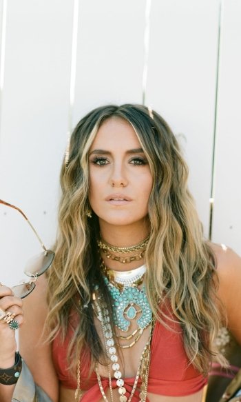 Phone wallpaper of a woman with long wavy hair, red top and layered turquoise-and-gold necklaces, holding sunglasses against a white fence — music-inspired, boho styling.