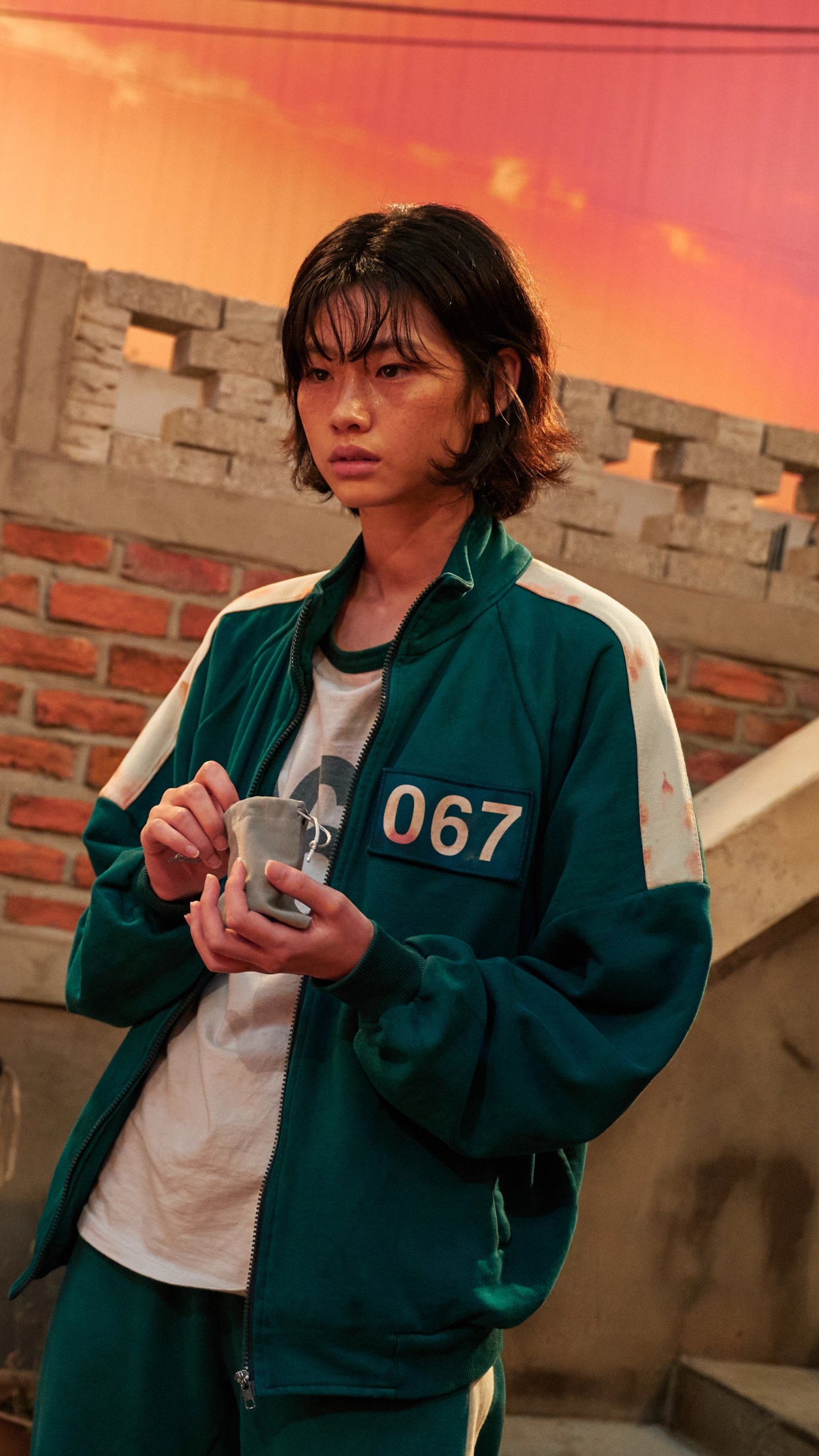 Phone wallpaper of Kang Sae-byeok from the TV show Squid Game, wearing a green 067 tracksuit and holding an object on a brick rooftop at sunset.