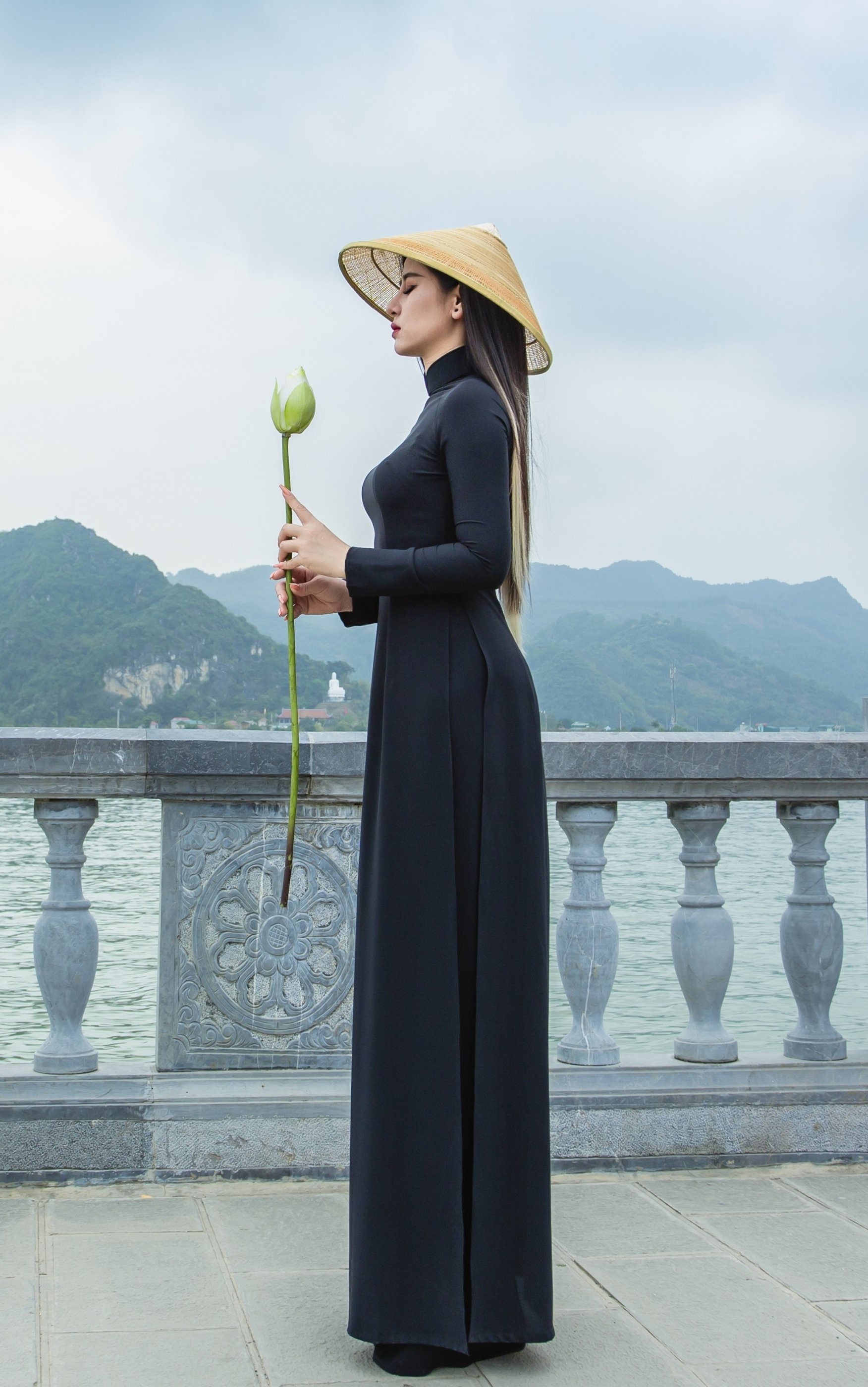 Phone wallpaper: a moody portrait of a woman in a long black traditional dress and conical hat, holding a lotus bud on a lakeside balustrade with misty mountains beyond.