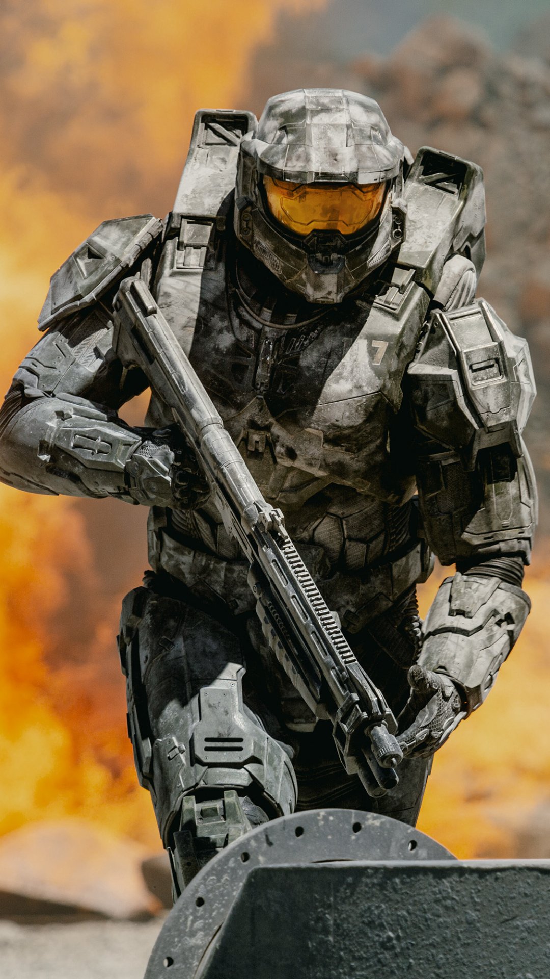 Phone wallpaper of Master Chief from the Halo TV show: armored Mjolnir suit with rifle and yellow visor, charging past a fiery orange explosion.