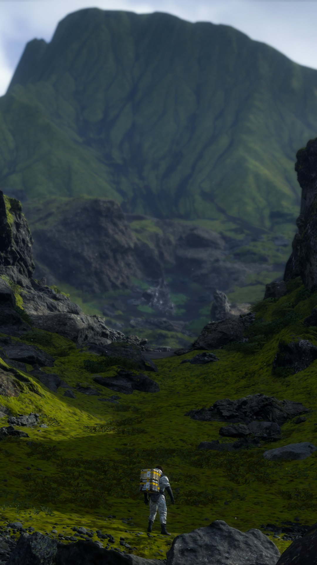 Phone wallpaper from the video game Death Stranding: a lone porter in cargo gear walks through a mossy valley toward a towering mountain beneath a pale sky.