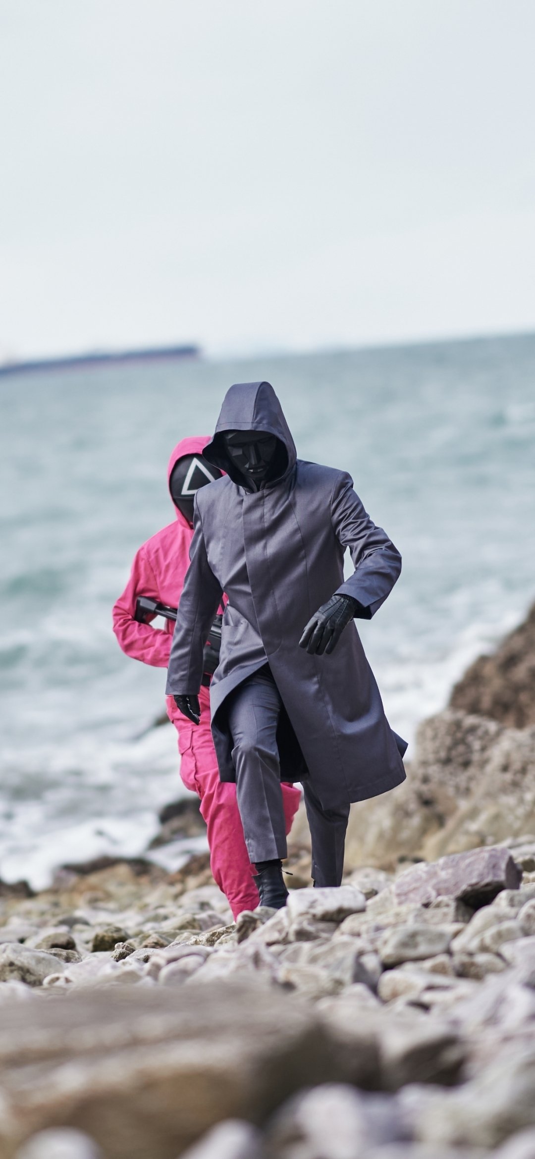 Phone wallpaper featuring masked characters from the TV show Squid Game walking along a rocky shore, designed for both iPhone and Android screens.