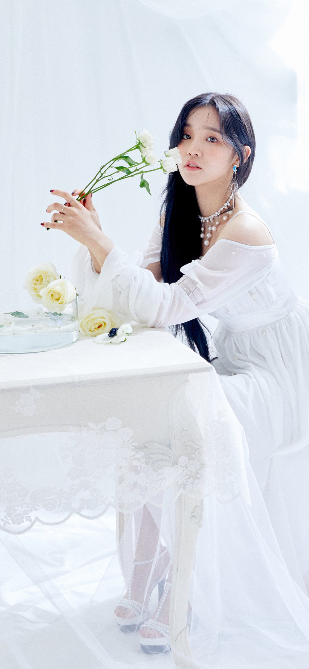 Phone wallpaper: K-pop singer in a flowing white dress holding white flowers at a lace-covered table — soft, music-themed portrait with ethereal, minimalist styling.