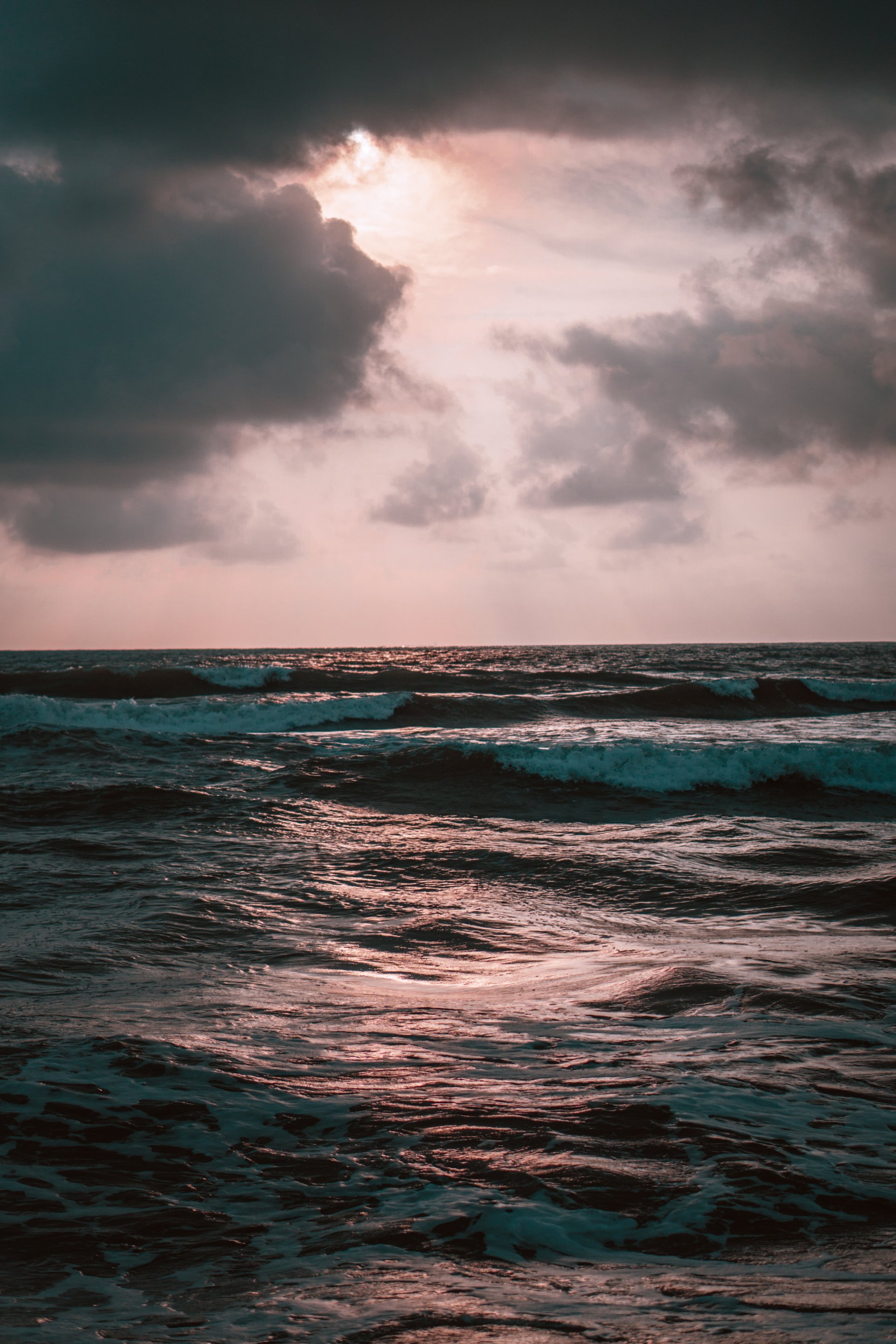 Phone wallpaper photograph of a beach at sunset: sunbeams breaking through dark clouds, warm light scattering across rippling ocean waves.