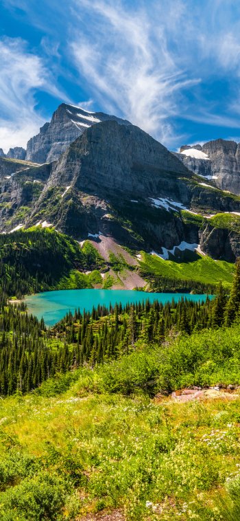 Vertical nature landscape iPhone wallpaper: turquoise alpine lake beneath towering rocky peaks, green wildflower meadow in foreground and wispy clouds across a bright blue sky.