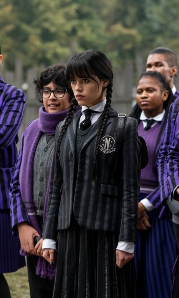 Phone wallpaper from the TV show Wednesday: a pale, dark‑haired girl with braids in a black striped school uniform stands solemnly among classmates in purple uniforms.