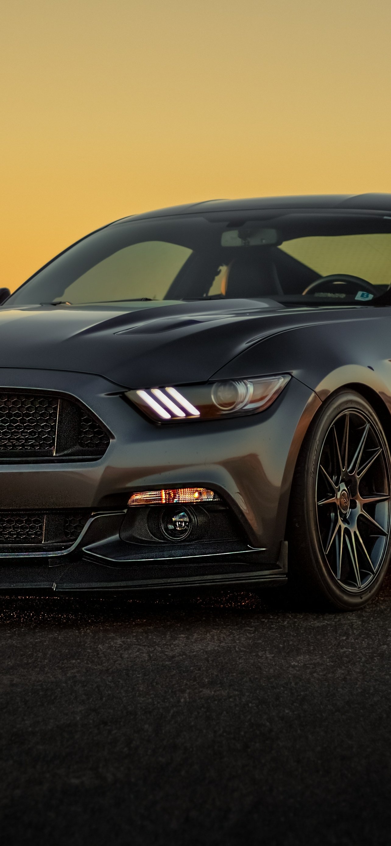 Phone wallpaper showing a sleek gray Ford Mustang vehicle at dusk, close-up front three-quarter view with illuminated headlights and alloy wheels.
