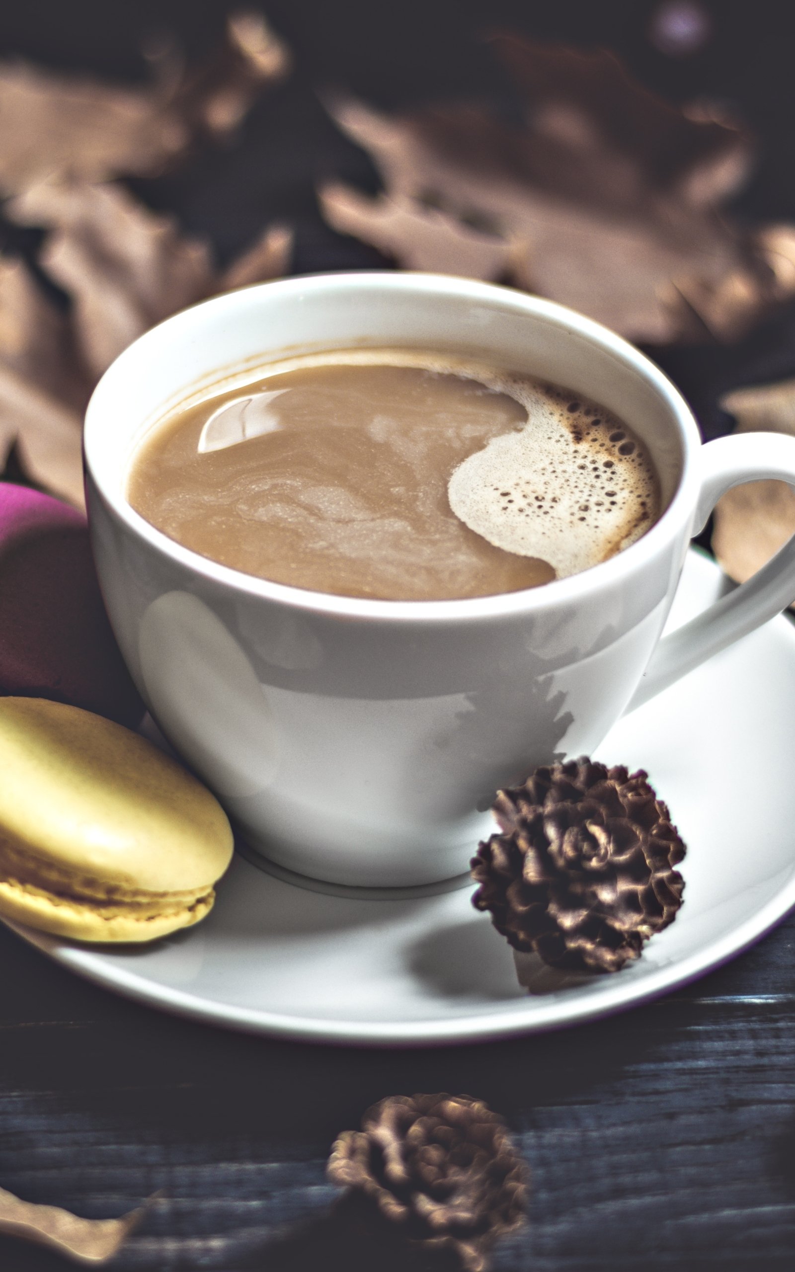 Phone wallpaper: creamy cup of coffee with froth on a saucer, a yellow macaron and a chocolate truffle on a dark, rustic background.