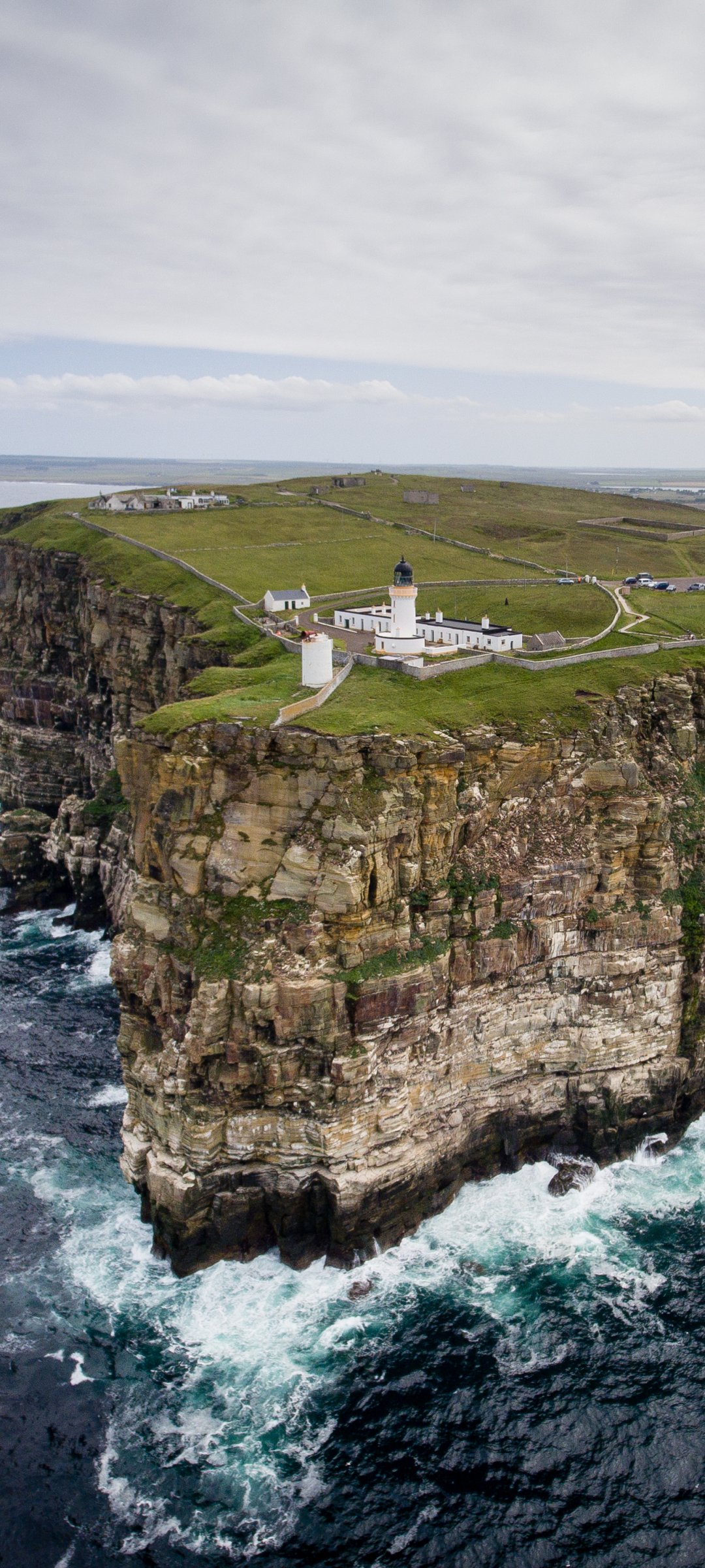 Phone wallpaper: man-made lighthouse perched on rugged coastal cliffs, waves crashing below and green fields stretching inland beneath a cloudy sky.
