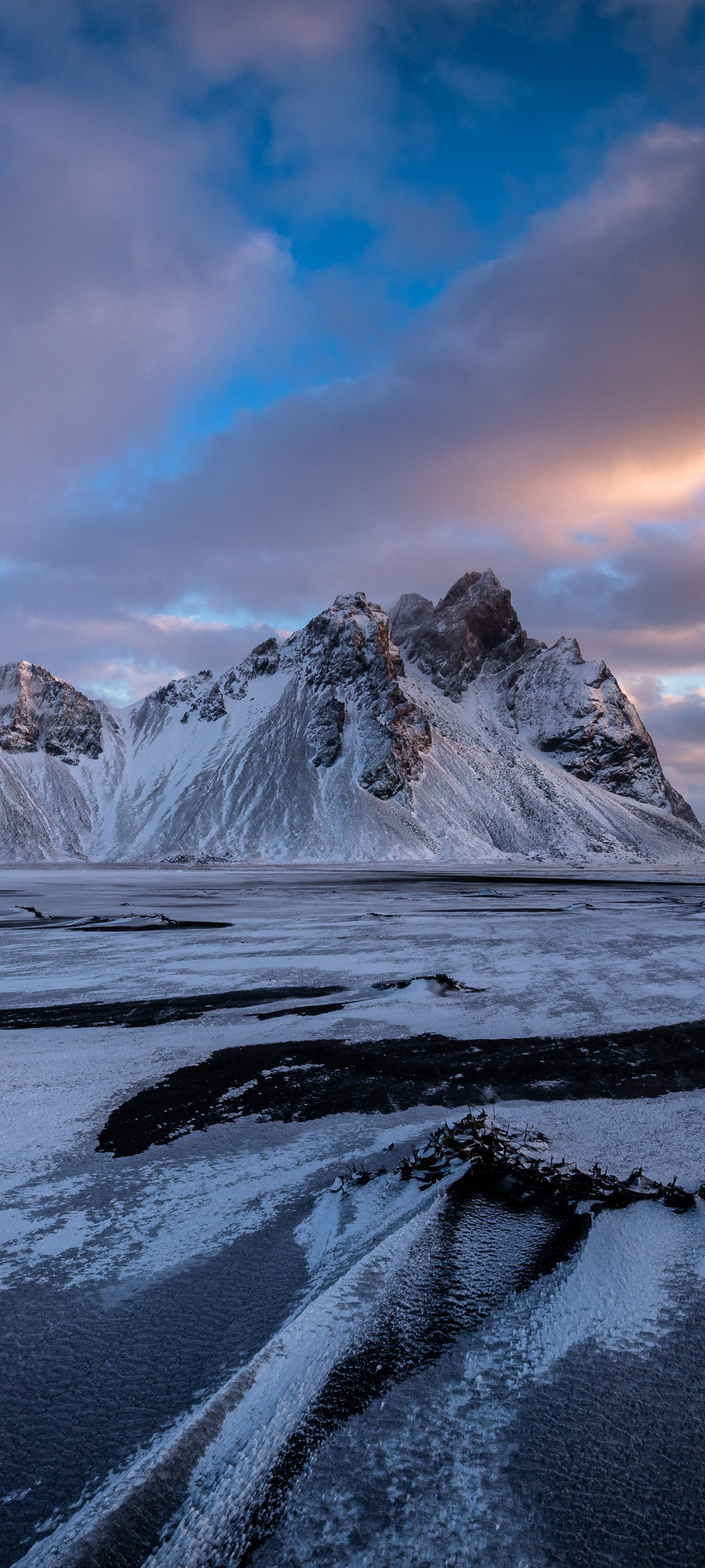 Vertical phone wallpaper of Vestrahorn: jagged snow-dusted peaks rising above a black-sand shoreline with icy tidal pools beneath a pastel blue and pink sky.