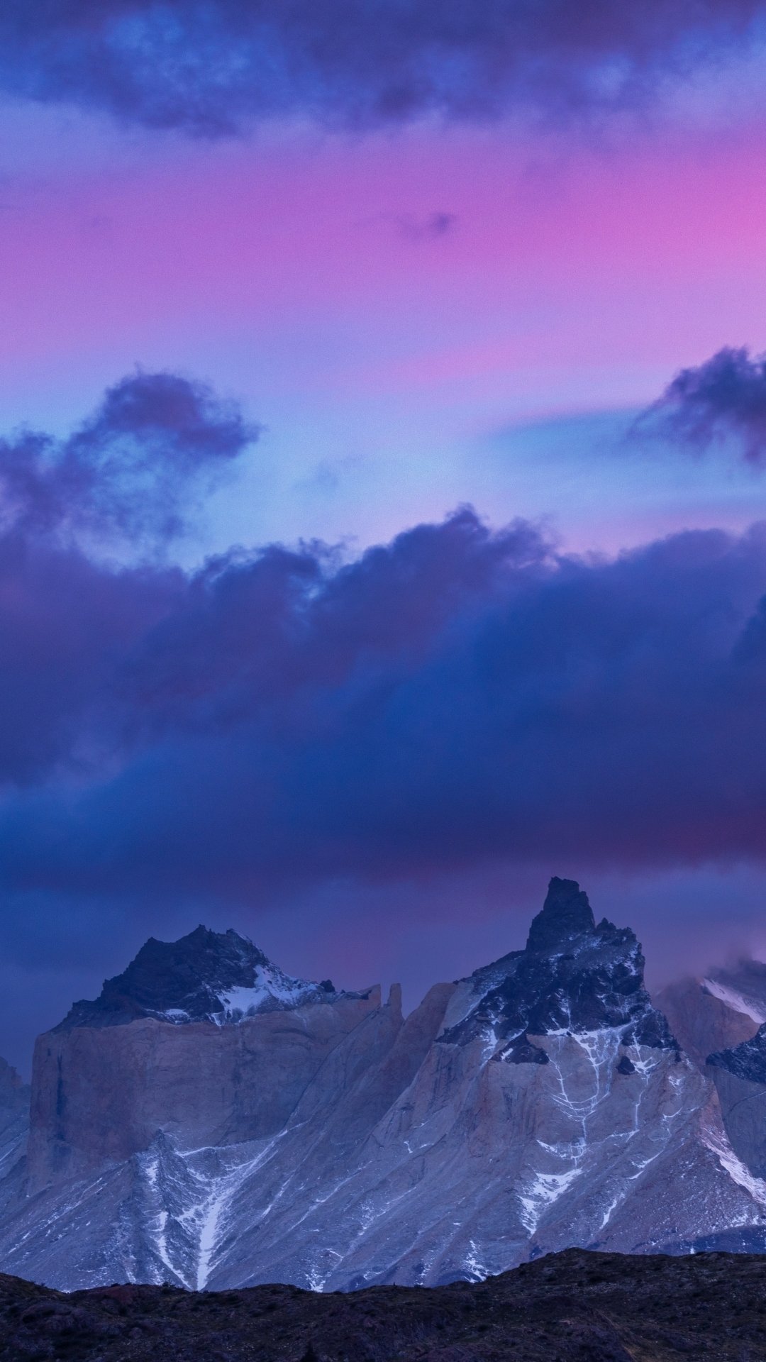 Phone wallpaper of Torres del Paine nature scene: jagged, snow-dusted peaks beneath a vivid pink and indigo sky with low clouds.