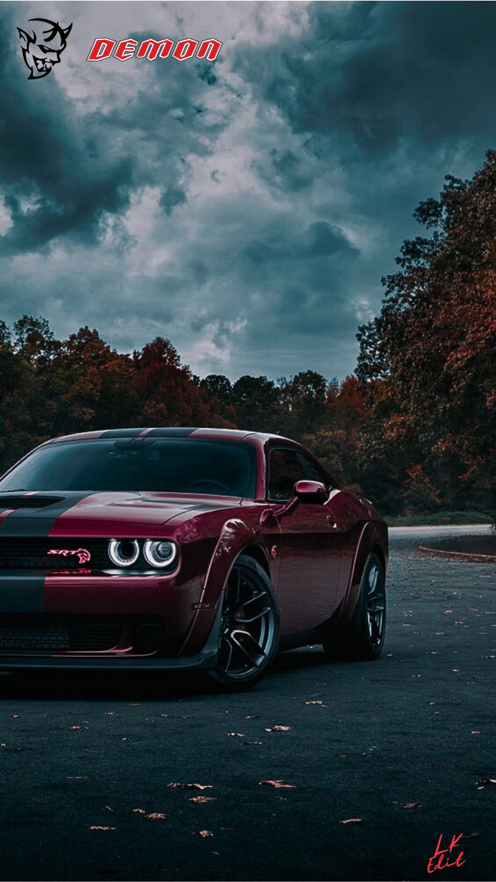 Phone wallpaper showing a dark red Dodge Challenger parked on an empty lot beneath dramatic storm clouds, 'Demon' logo top-left and small artist signature bottom-right.