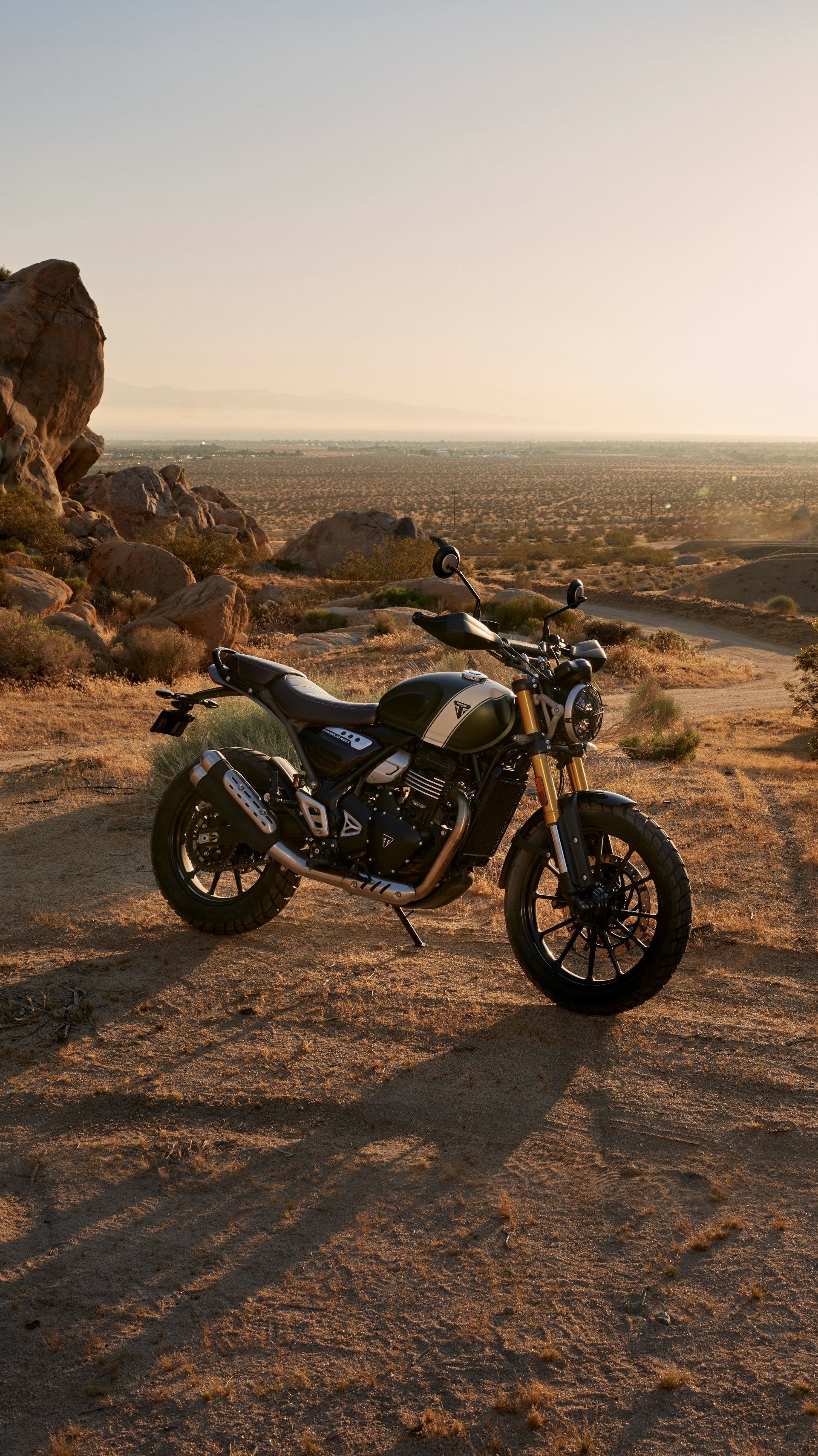 A Triumph Scrambler 400 X stands on a rocky landscape, basking in the warm glow of sunset, showcasing its rugged design against a scenic backdrop. A great phone wallpaper choice.