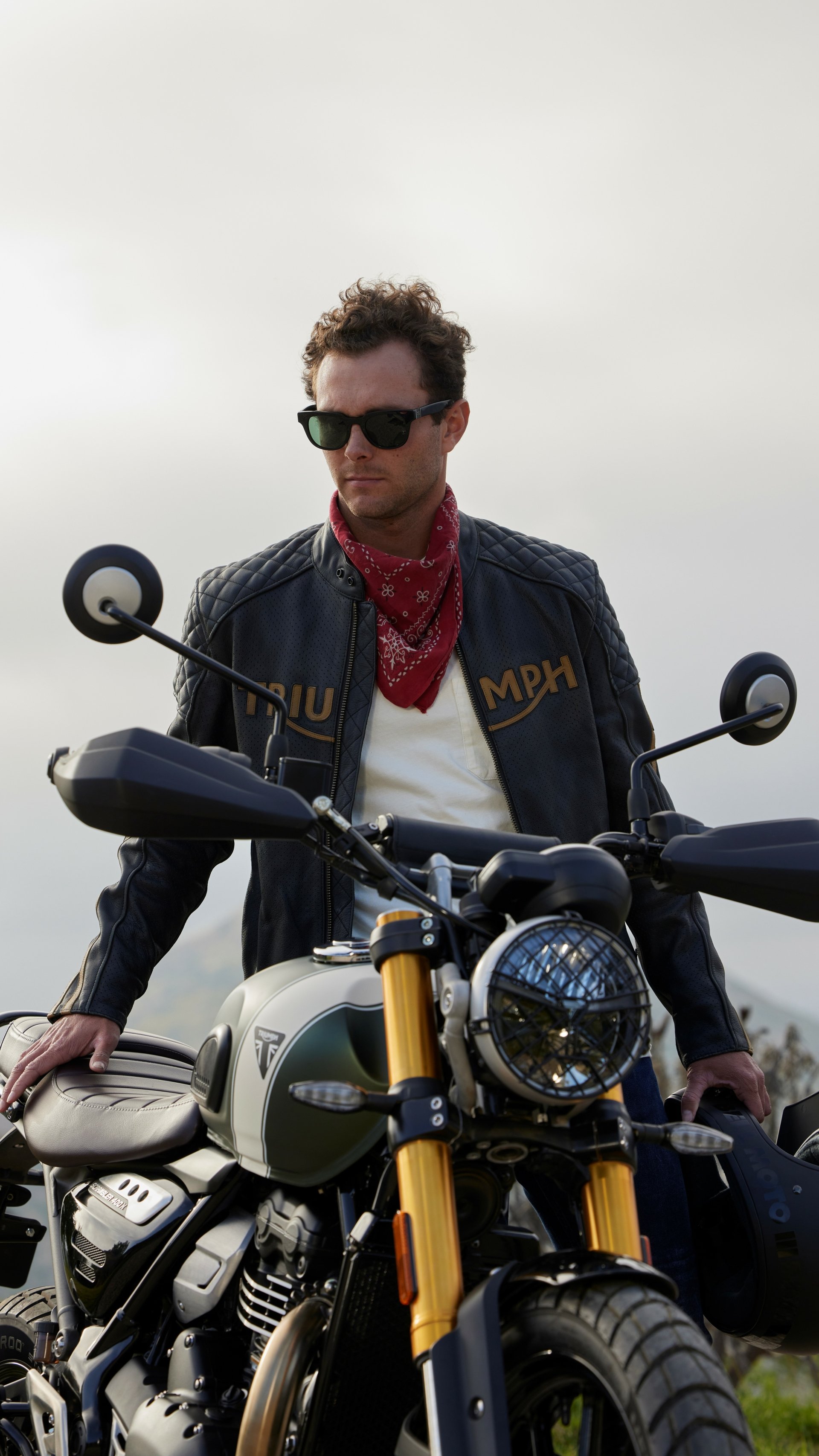 A stylish rider in sunglasses and a bandana stands beside a Triumph Scrambler 400 X, set against a scenic backdrop, creating an adventurous phone wallpaper.