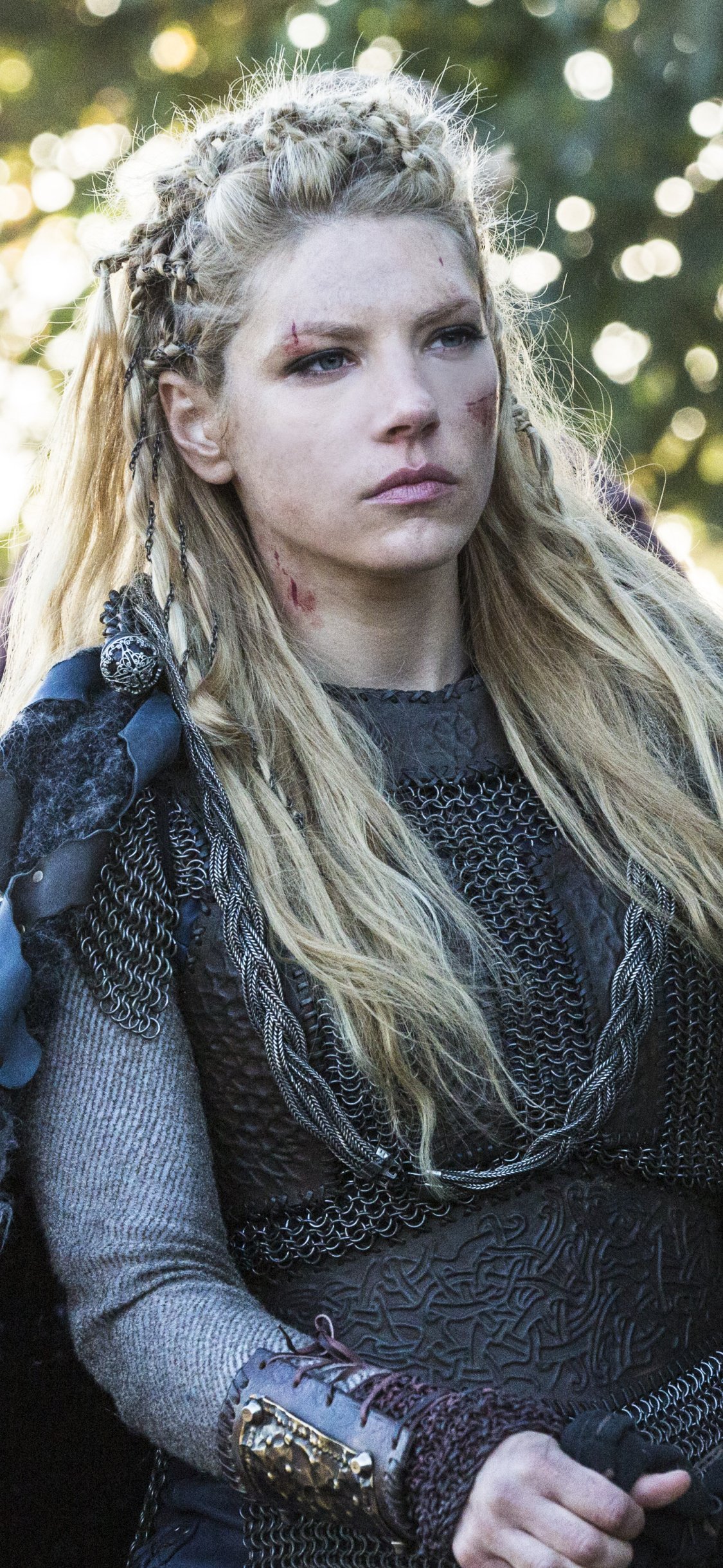 Phone wallpaper: a fierce shield‑maiden from TV show Vikings (2013) — braided blonde hair, scarred cheek, layered leather armor and a pensive, battle‑ready expression.