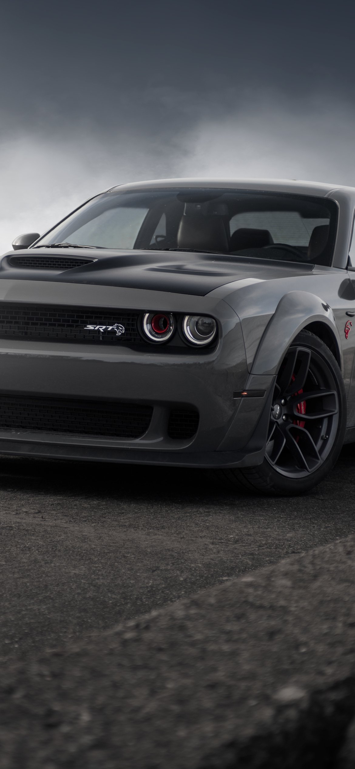 Vertical phone wallpaper: close-up of a gray Dodge Challenger SRT Demon on asphalt beneath a dramatic cloudy sky.
