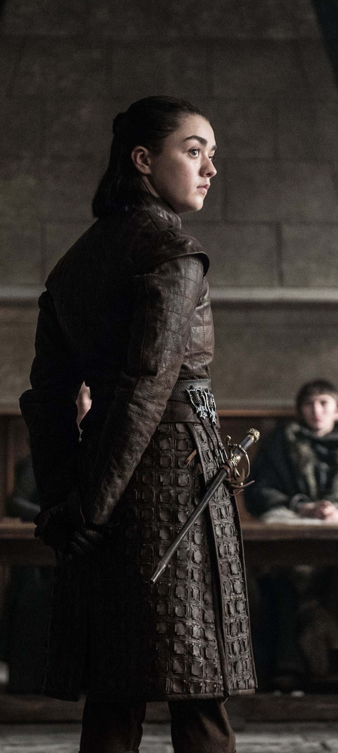 Game of Thrones phone wallpaper depicting a young woman in leather armor, hand on a sword at her hip, standing in a dim stone hall and looking to the side.