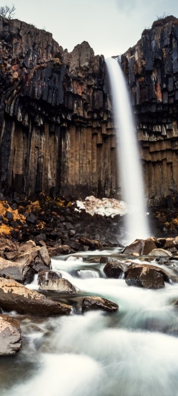 Phone nature wallpaper of Svartifoss: a narrow waterfall plunging between dark hexagonal basalt columns into a misty, rock-strewn river.